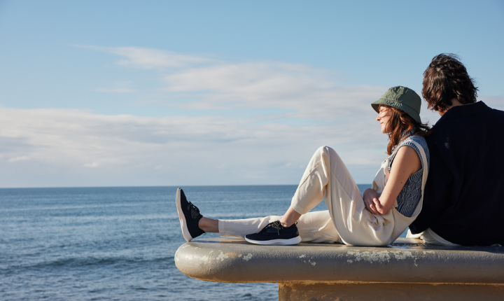 Woman, sitting next to man on bench that overlooks the sea, wearing cream overalls and Vessi Everyday Move Sneakers.